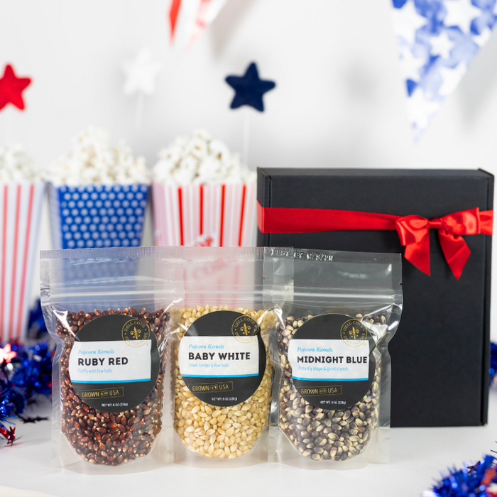 Black gift box tied closed with red ribbon next to 3 half pound popcorn bags, Ruby Red, Baby White and Midnight Blue with large black bowl filled with popped popcorn on red white and blue background - Dell Cove Spices